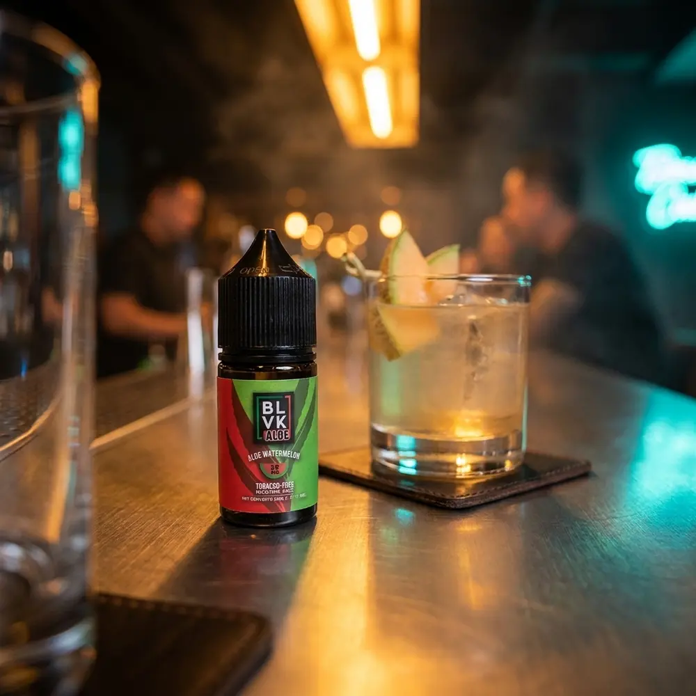 Frasco 30ml Blvk Unicorn Salt Aloe Watermelon apoiado em balcão metálico de lounge, luzes cálidas e copo de coquetel ao fundo - Vapor Gringo