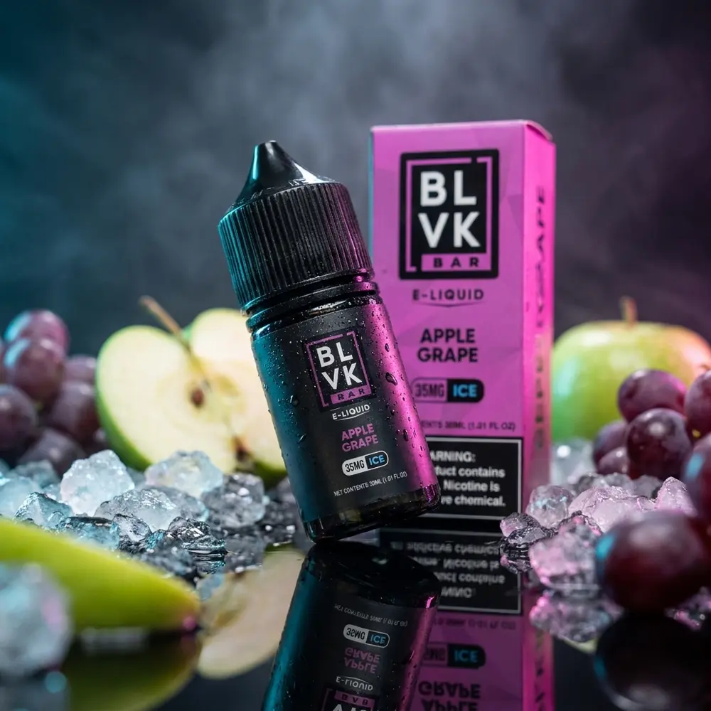 Garrafa BLVK Unicorn Salt Apple Grape Ice inclinada sobre base preta com gelo, fatias de maçã e reflexos azuis - Vapor Gringo