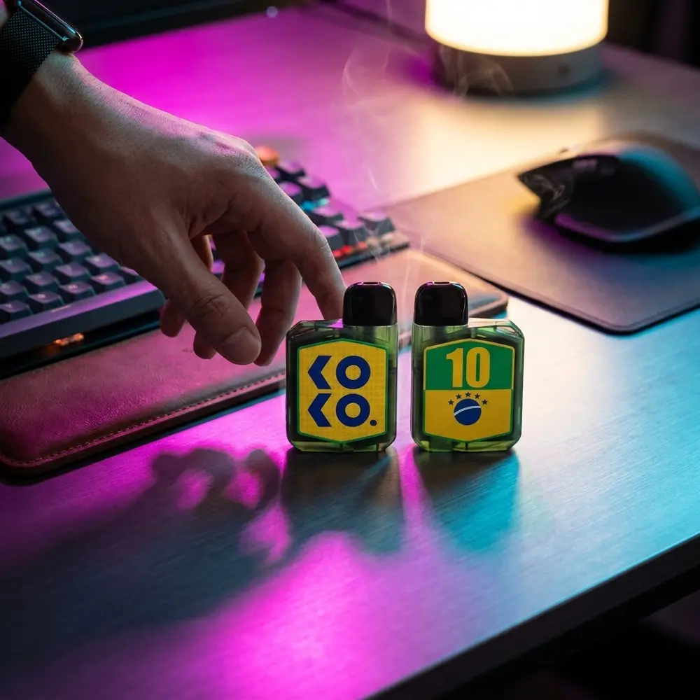 Kit Pod Caliburn Koko Prime Vision repousando em mesa gamer RGB, teclado mecanico e luzes neon ao fundo - Vapor Gringo