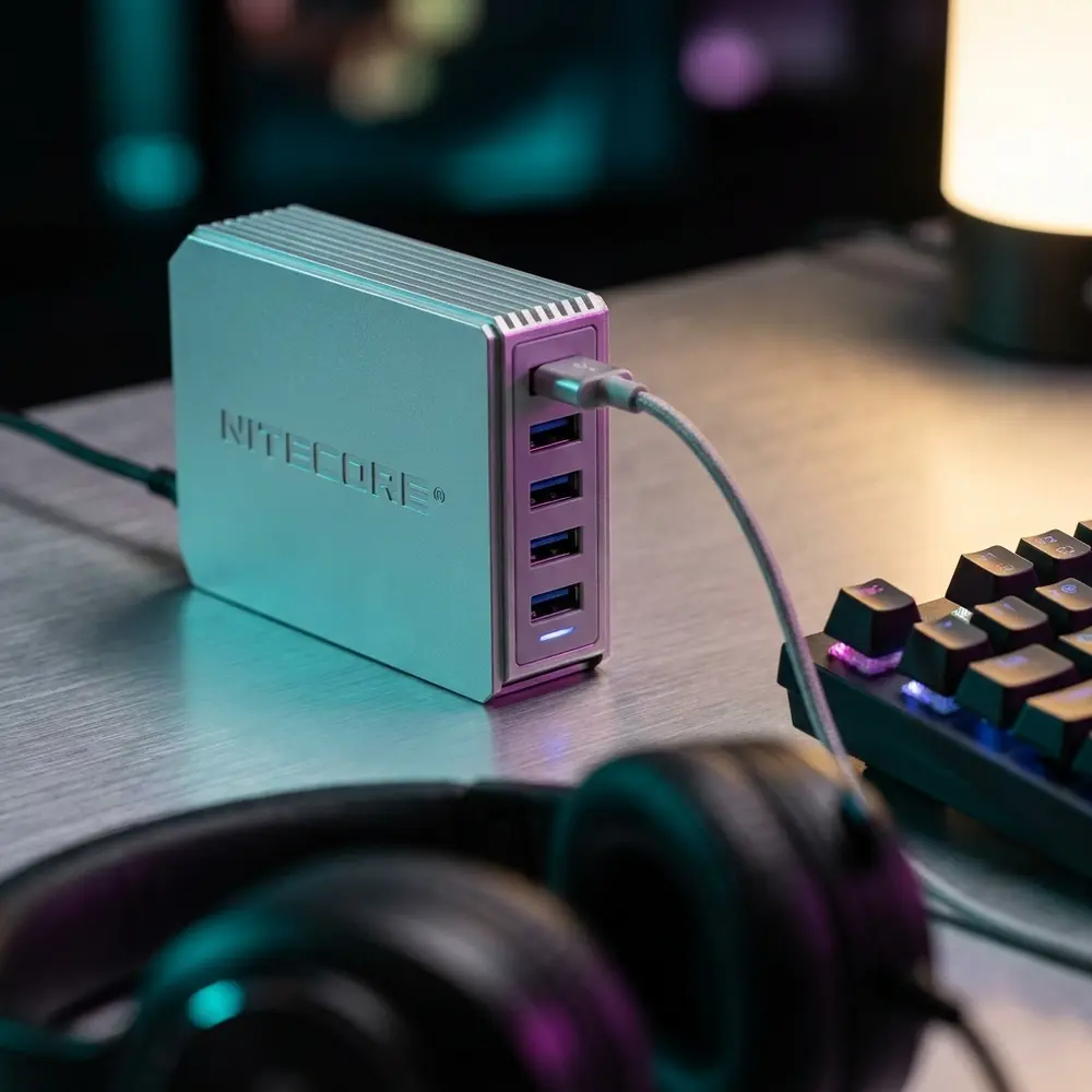 Carregador UA55 Nitecore em mesa gamer com teclado RGB, fone desfocado e luzes noturnas urbanas - Vapor Gringo