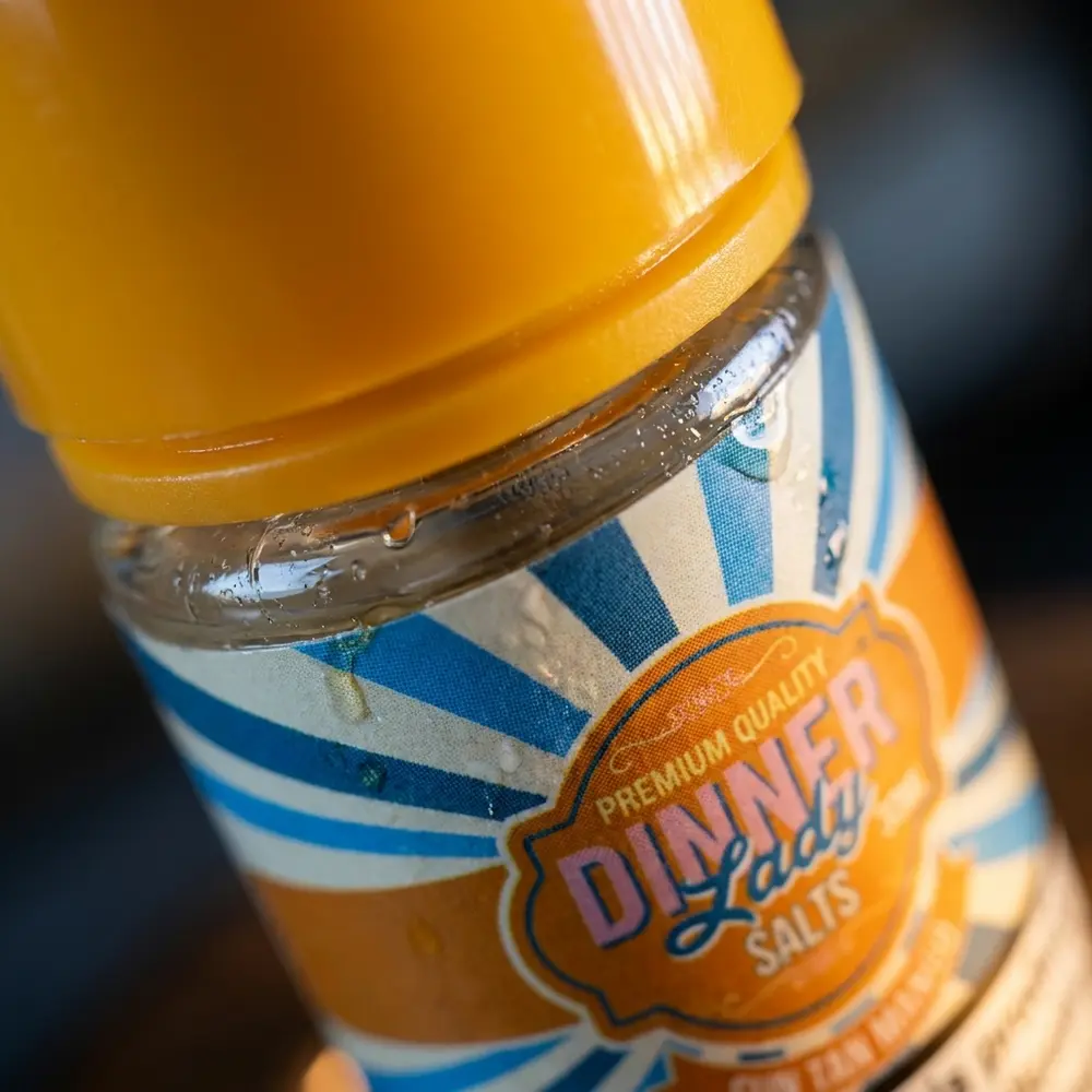 Close-up do rótulo e bico do frasco Dinner Lady Sun Tan Mango com gotas de condensação e reflexos detalhados - Vapor Gringo