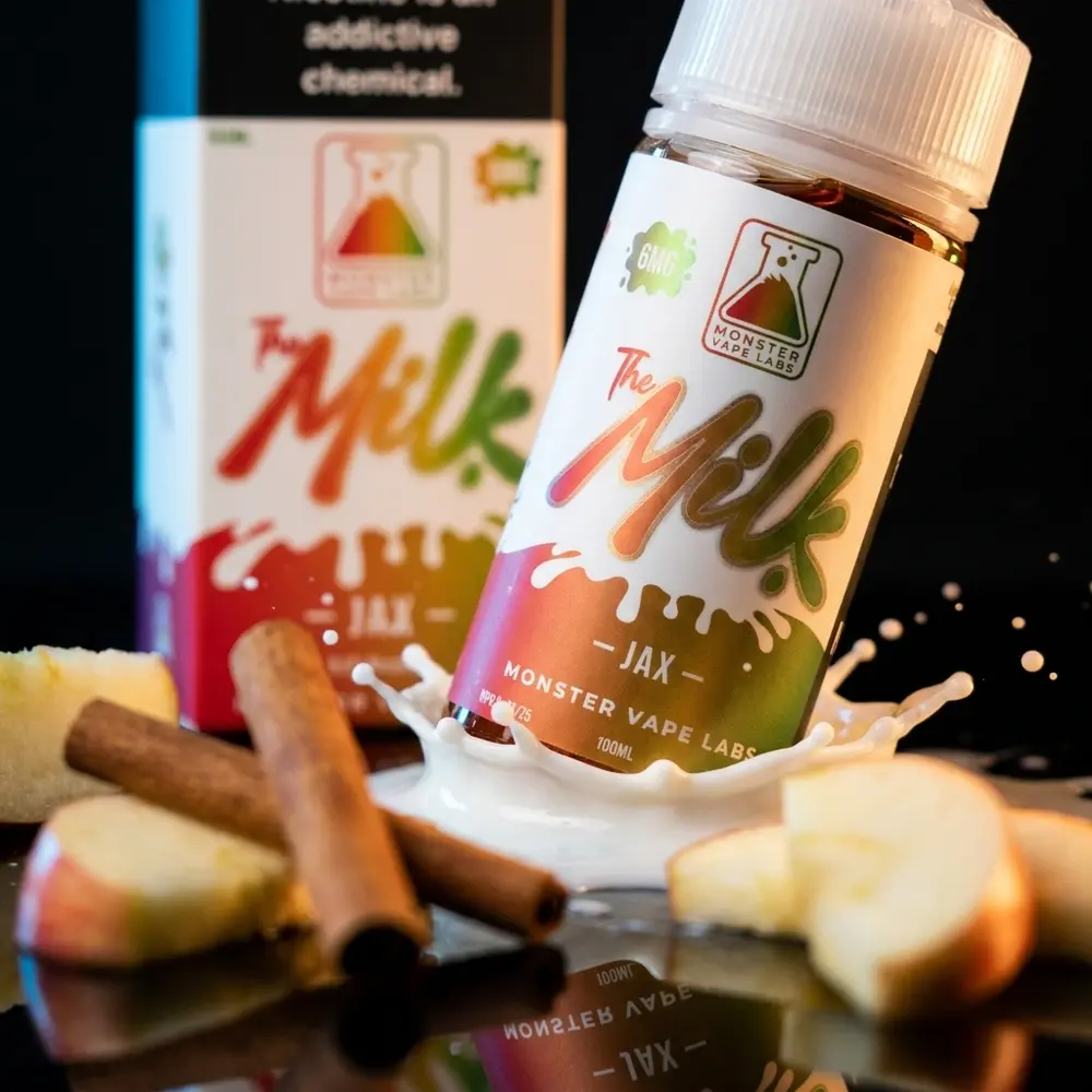 Frasco Liquido Jam Monster The Milk Jax inclinado sobre acrilico preto com gotas de leite, fatia de maca e canela - Vapor Gringo