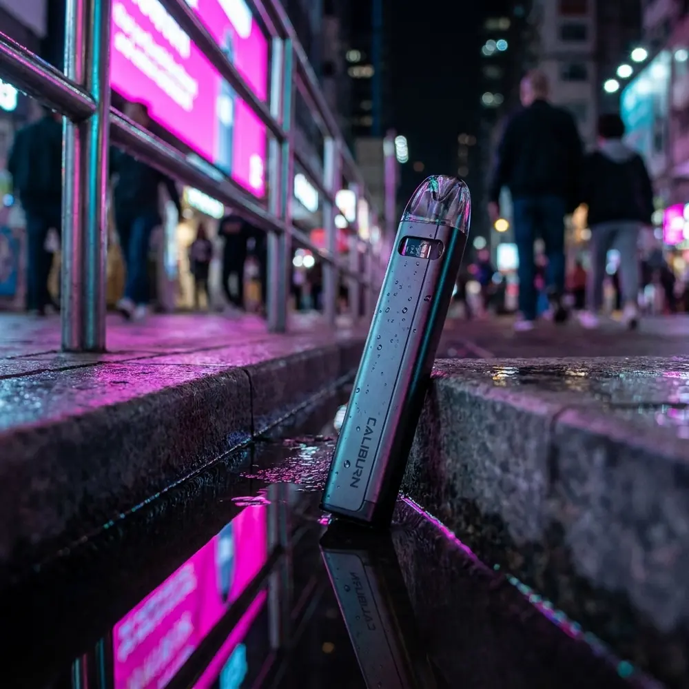 Kit Pod Caliburn A2s apoiado em guia molhada na rua à noite com luzes neon refletidas e gotas visíveis - Vapor Gringo