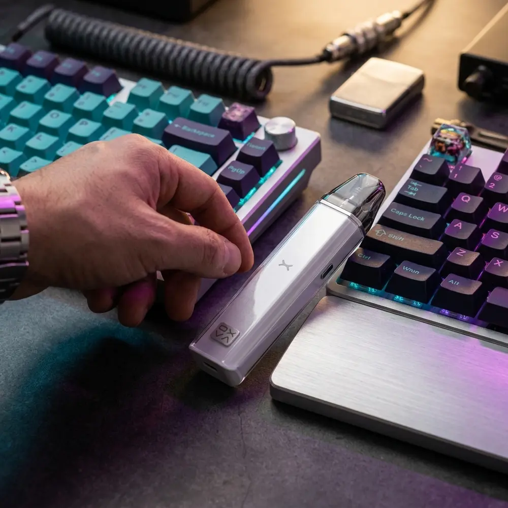 Kit Pod Xlim Crystal - Oxva apoiado em teclado RGB, ambiente gaming urbano com reflexos e mao parcial - Vapor Gringo