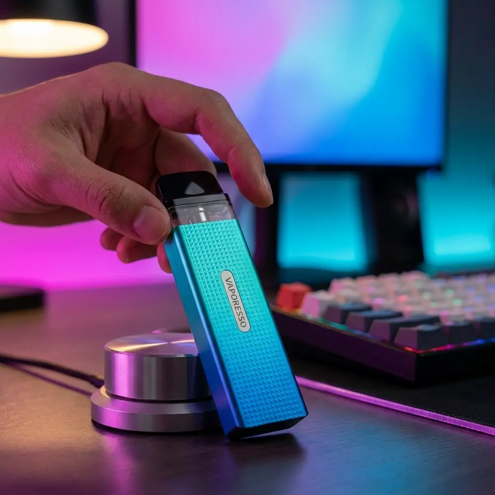 Vaporesso Xros Mini ao lado de teclado RGB em mesa gamer, luzes magenta e ciano refletindo no chassi - Vapor Gringo