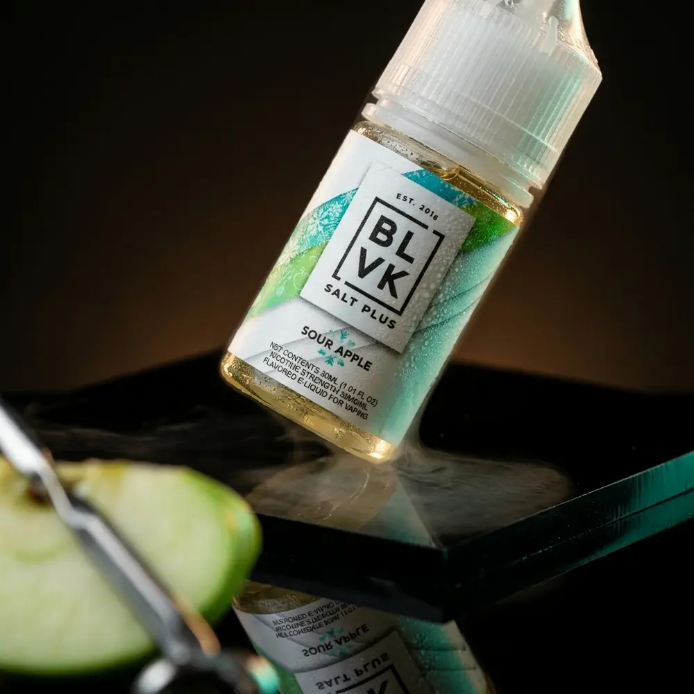 Frasco BLVK Salt Plus Sour Apple Ice inclinado sobre base acrílica preta com reflexos verdes e gelo condensado - Vapor Gringo