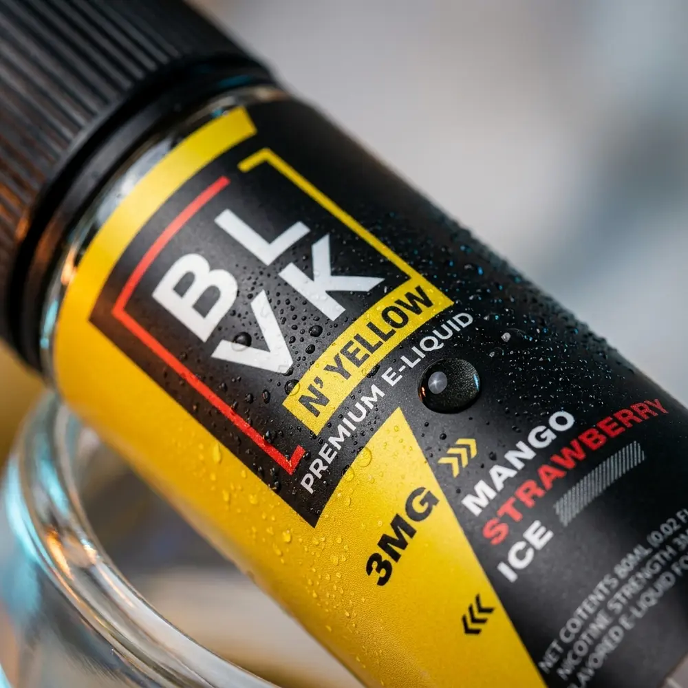 Close-up do rótulo e bocal do Líquido Blvk Unicorn Mango Strawberry Ice Yellow com gotas de condensação e textura do vidro - Vapor Gringo