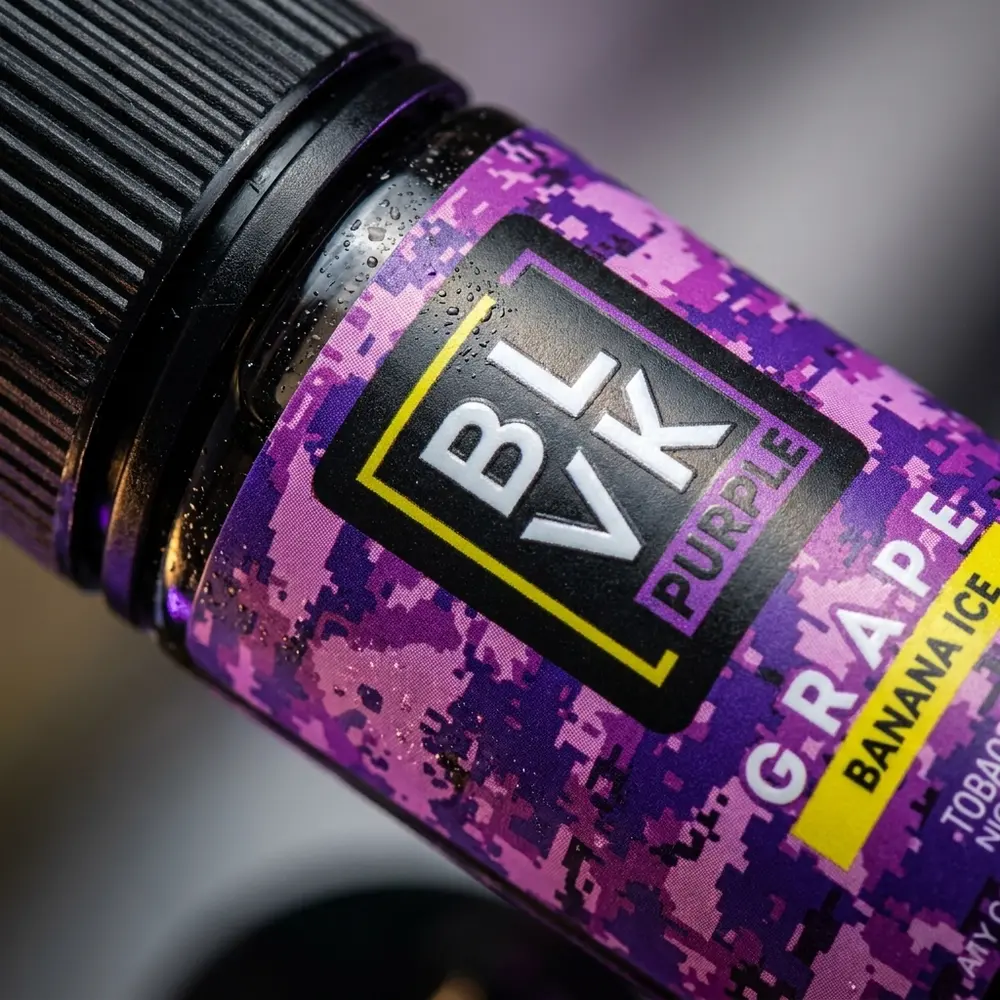 Close-up do rótulo e bico do Líquido Blvk Unicorn Salt Grape Banana Ice com gotas e micro-reflexos - Vapor Gringo