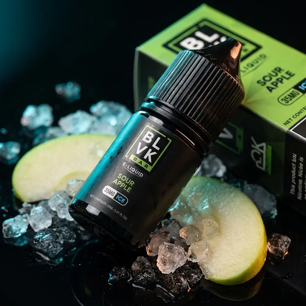 Garrafa do Líquido BLVK Unicorn Sour Apple Ice sobre acrílico com fatias de maçã e gelo, iluminação contrastante - Vapor Gringo