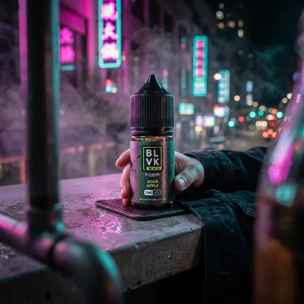 Líquido BLVK Unicorn Sour Apple Ice apoiado num balcão urbano molhado com luzes neon e itens de lifestyle noturno - Vapor Gringo