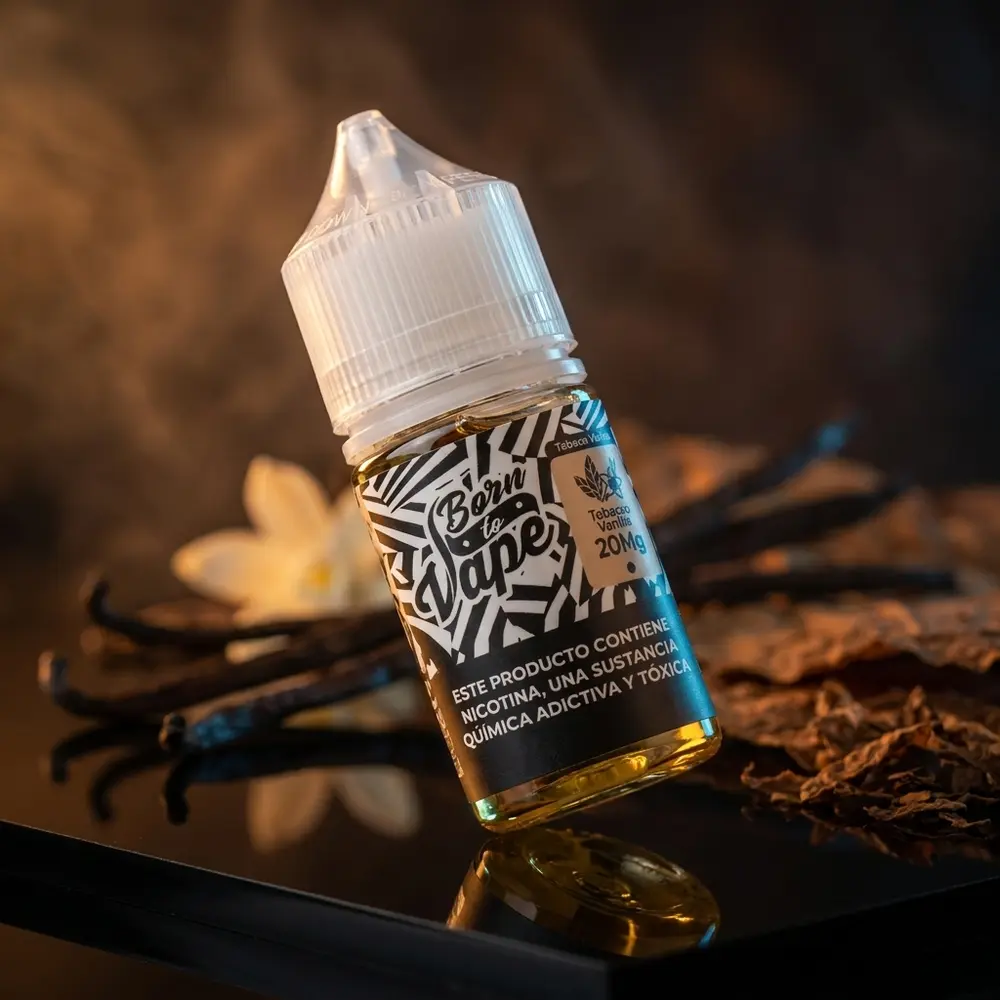 Frasco Born to Vape Salt Tobacco Vanilla inclinado sobre acrilico preto brilhante, luz ambar e vagens de baunilha desfocadas ao fundo - Vapor Gringo
