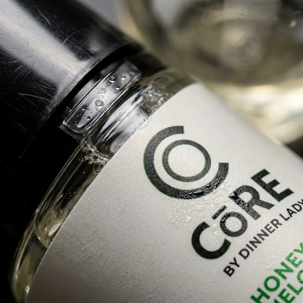 Close-up do rótulo e bico da garrafa Líquido Core Honeydew, gotas de condensação e textura impressa - Vapor Gringo