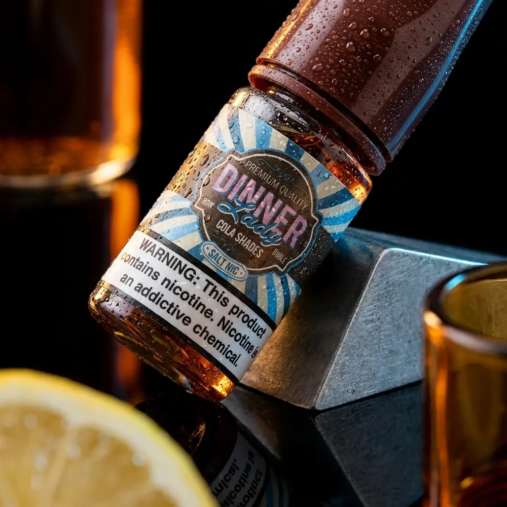 Frasco do Líquido Dinner Lady Salt Cola Shades 30ml inclinado sobre acrílico preto com gotas e reflexos âmbar - Vapor Gringo