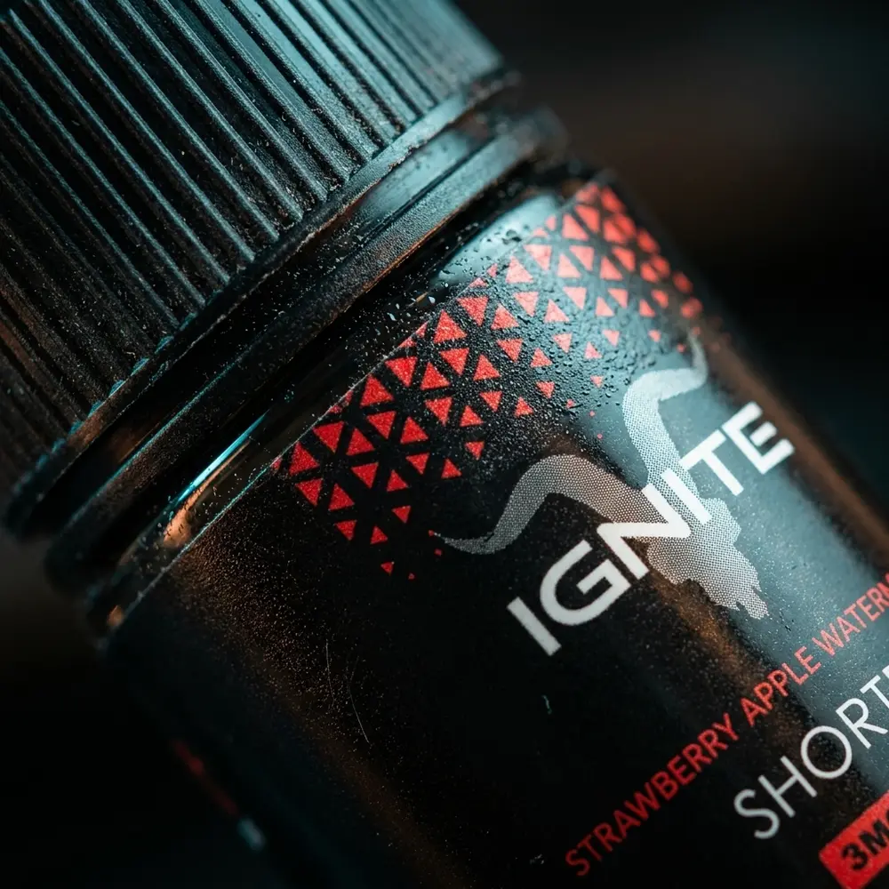 Close-up do bocal e rótulo do Líquido Ignite Strawberry Apple Watermelon Ice com gotas de condensação e reflexos metálicos. - Vapor Gringo