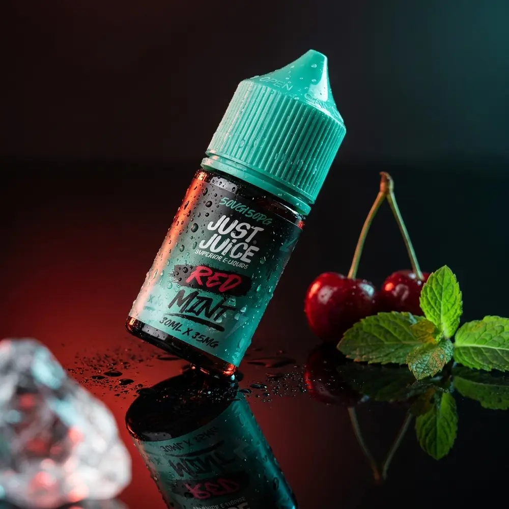 Garrafa do Líquido Just Juice Salt Red Mint inclinada sobre acrílico preto com condensação, cerejas e folhas de menta - Vapor Gringo