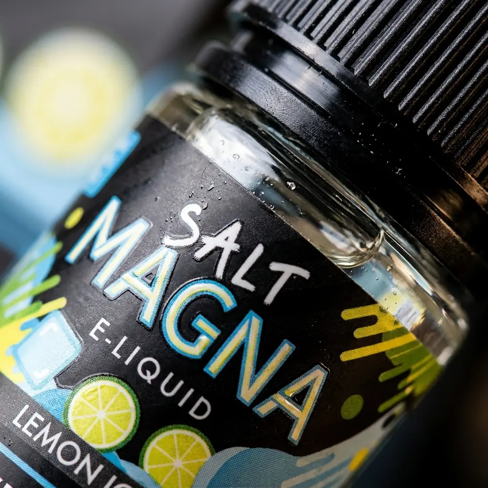 Detalhe macro do rótulo e bico do Liquido Magna Lemon Ice com gotas, textura do vidro e reflexos. - Vapor Gringo