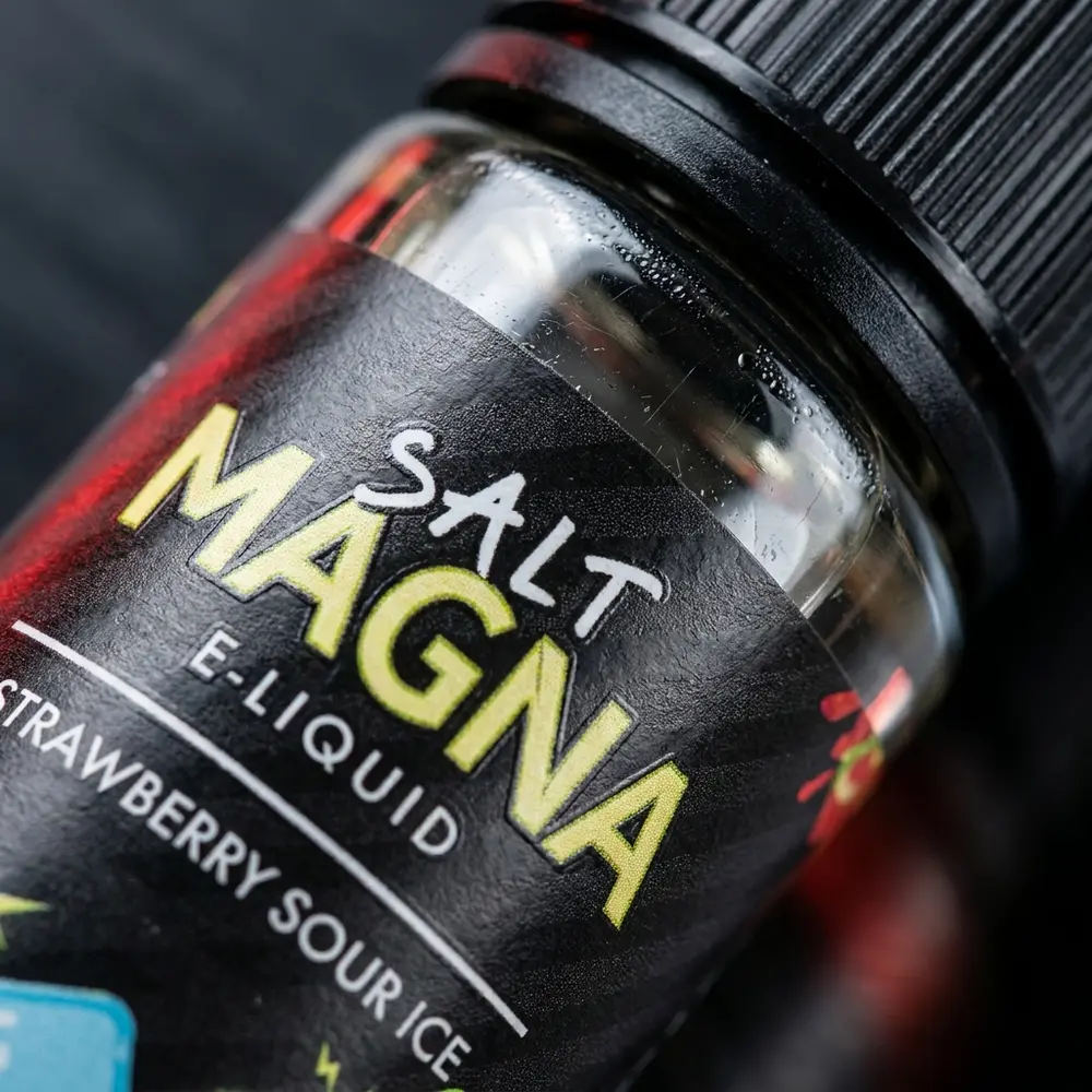Detalhe do rótulo e bico do Líquido Magna Strawberry Sour Ice com condensação, textura do vidro e reflexos metálicos - Vapor Gringo