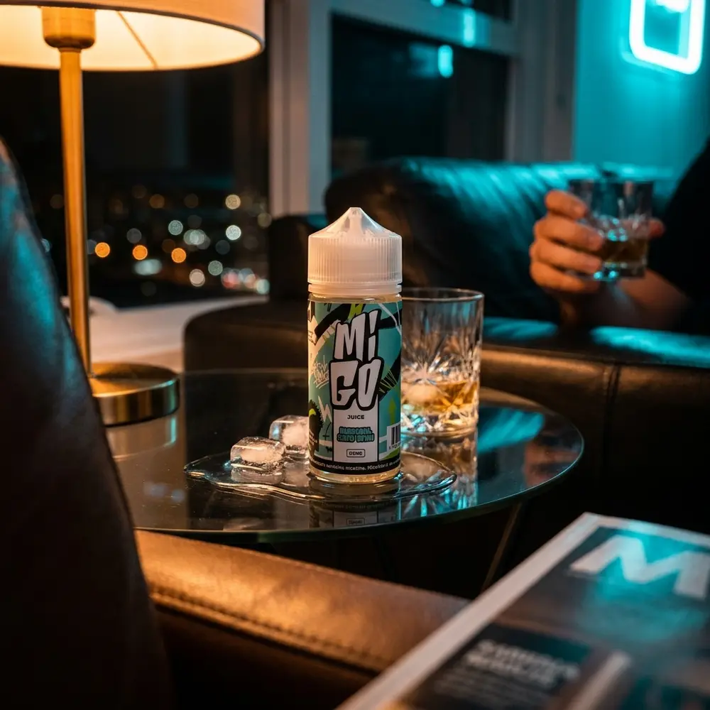 Liquido Migo Menthol Zero Grau apoiado em mesa de lounge luxuoso com gelo, reflexos âmbar e cyan e bokeh urbano - Vapor Gringo