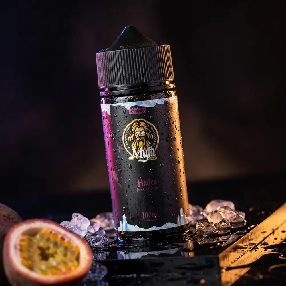 Líquido Myth Hades Passion Fruit Ice 100ml sobre base acrílica preta molhada com gotas e maracujá ao fundo - Vapor Gringo