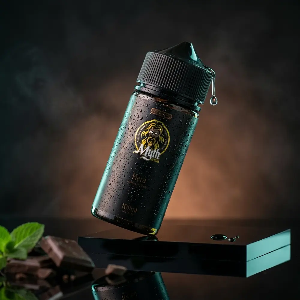 Frasco do Líquido Myth Hera 100ml Chocolate Mint sobre acrílico preto com gotas, pedaços de chocolate e folha de menta. - Vapor Gringo