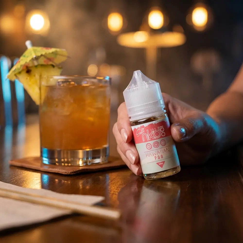 Frasco Liquido Naked 100 Salt Hawaiian Pog ICE no balcão de um lounge elegante, coquetel e luzes quentes ao fundo - Vapor Gringo