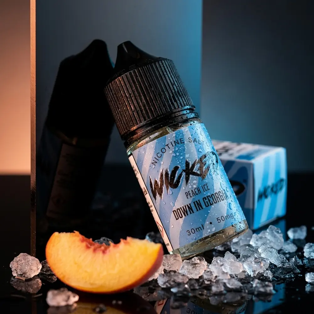 Frasco Wicked Salt Peach Ice inclinado sobre acrílico escuro com gelo e fatia de pêssego, estúdio premium. - Vapor Gringo