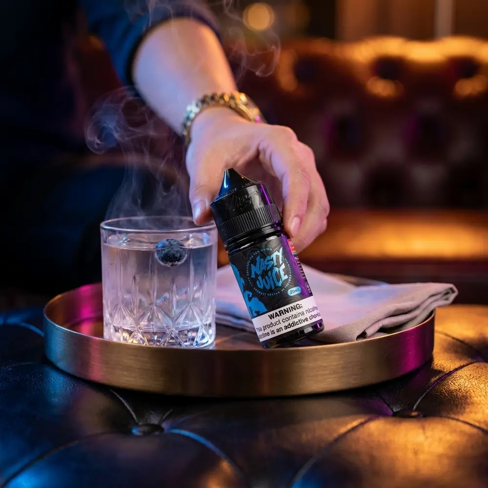 Garrafa Nasty Juice Salt Blueberry em bandeja de bronze no lounge luxuoso, luzes azul e ambar ao fundo - Vapor Gringo