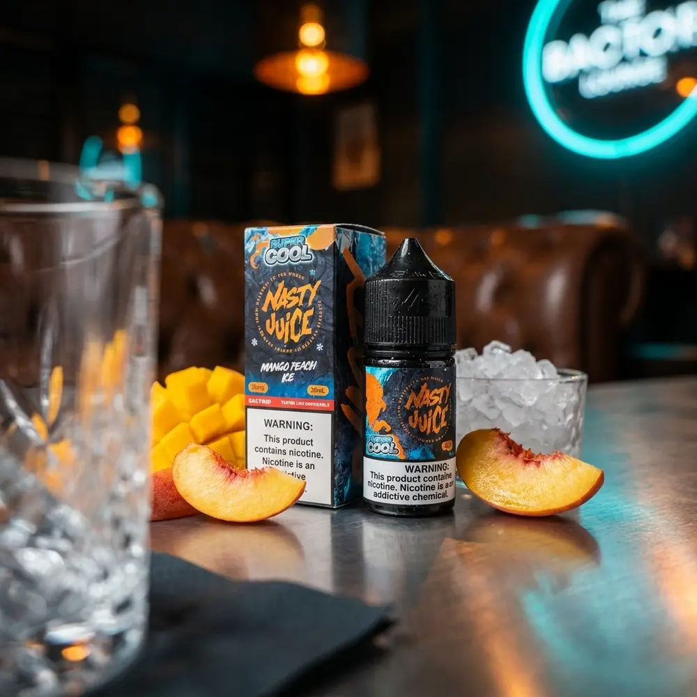 Frasco Nasty Juice Salt Super Cool Mango Peach Ice sobre mesa de lounge, fatias de manga e pêssego com luzes neon ao fundo - Vapor Gringo