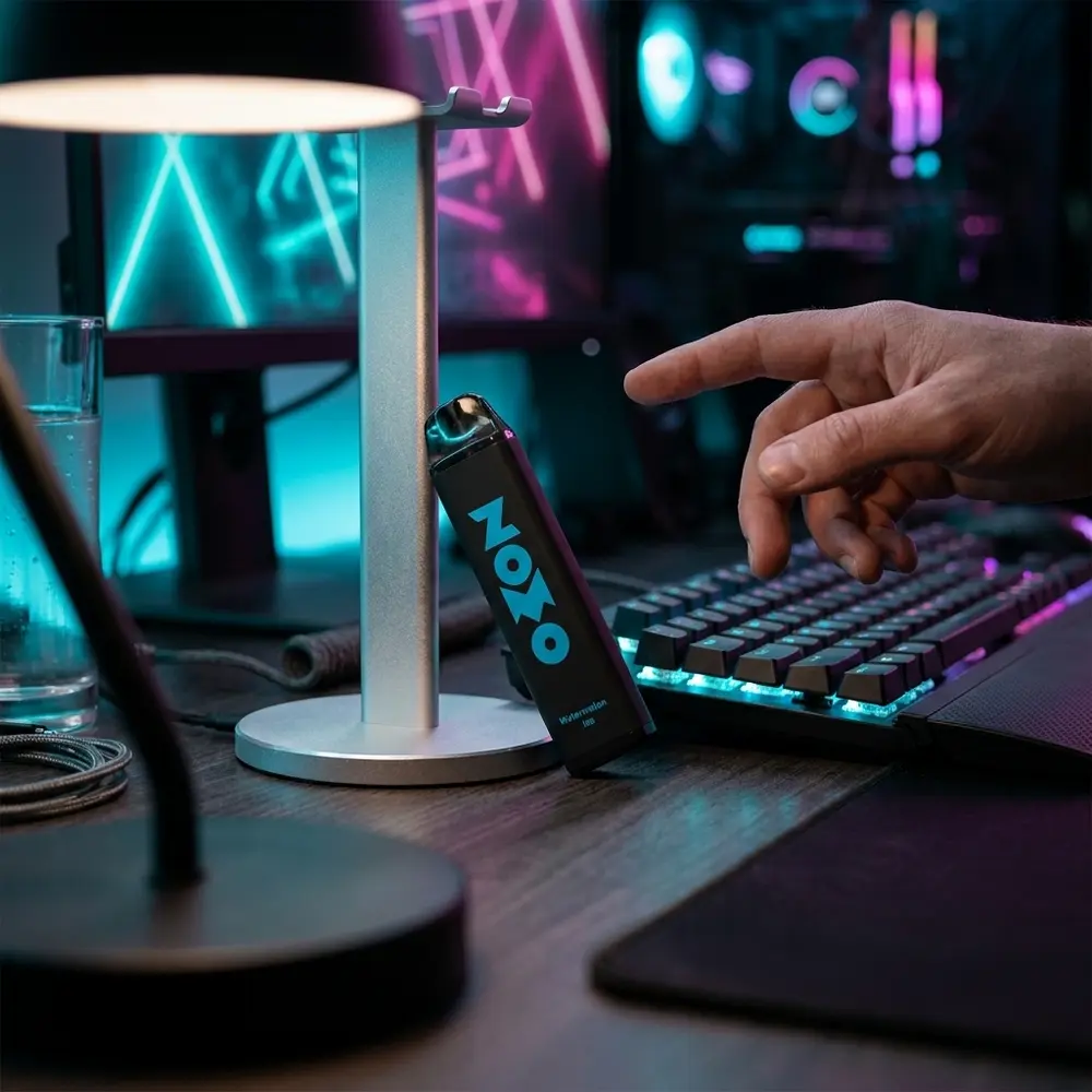 Pod descartavel Zomo Melon Ice 800 puffs apoiado em setup gamer RGB com teclado mecanico e luzes neon ao fundo - Vapor Gringo