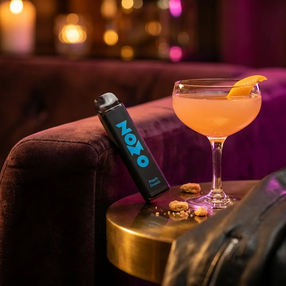 Pod descartavel Zomo Party Peach Crunch apoiado em sofa de veludo com cocktail de pessego e luzes de lounge - Vapor Gringo