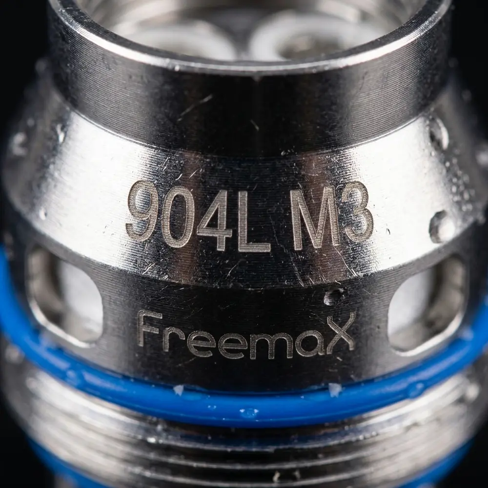 Detalhe macro da marca e textura da Resistência Coil 904L M Freemax com gotas e reflexos metálicos - Vapor Gringo