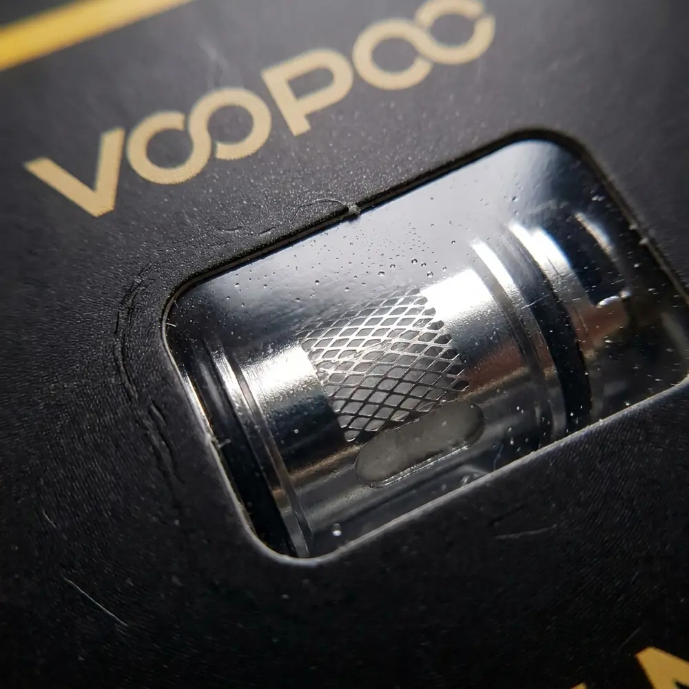 Detalhe macro da Resistência Coil MT Voopoo mostrando texturas, janela plástica, malha metálica e reflexos - Vapor Gringo