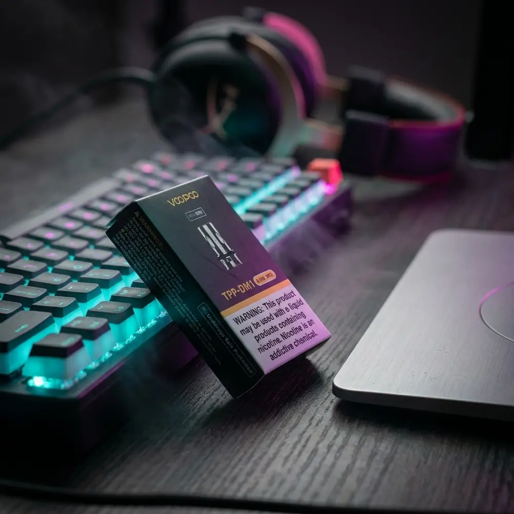 Caixa da resistencia Voopoo TPP-DM1 0,15Ω apoiada em mesa gamer RGB entre teclado e headset, luzes coloridas - Vapor Gringo