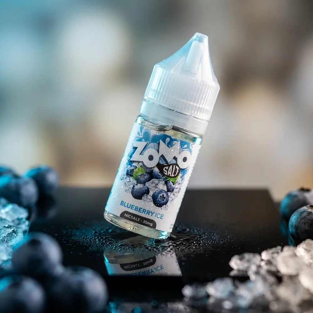 Líquido Zomo Salt Blueberry Ice em frasco sobre acrílico preto, iluminação azul gelada e gotas de condensação - Vapor Gringo
