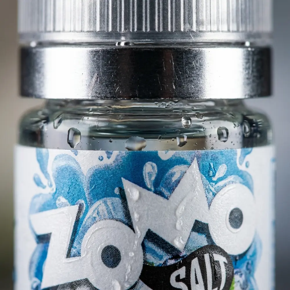 Detalhe do bico e rótulo do Líquido Zomo Salt Blueberry Ice com gotas de condensação e reflexos metálicos - Vapor Gringo